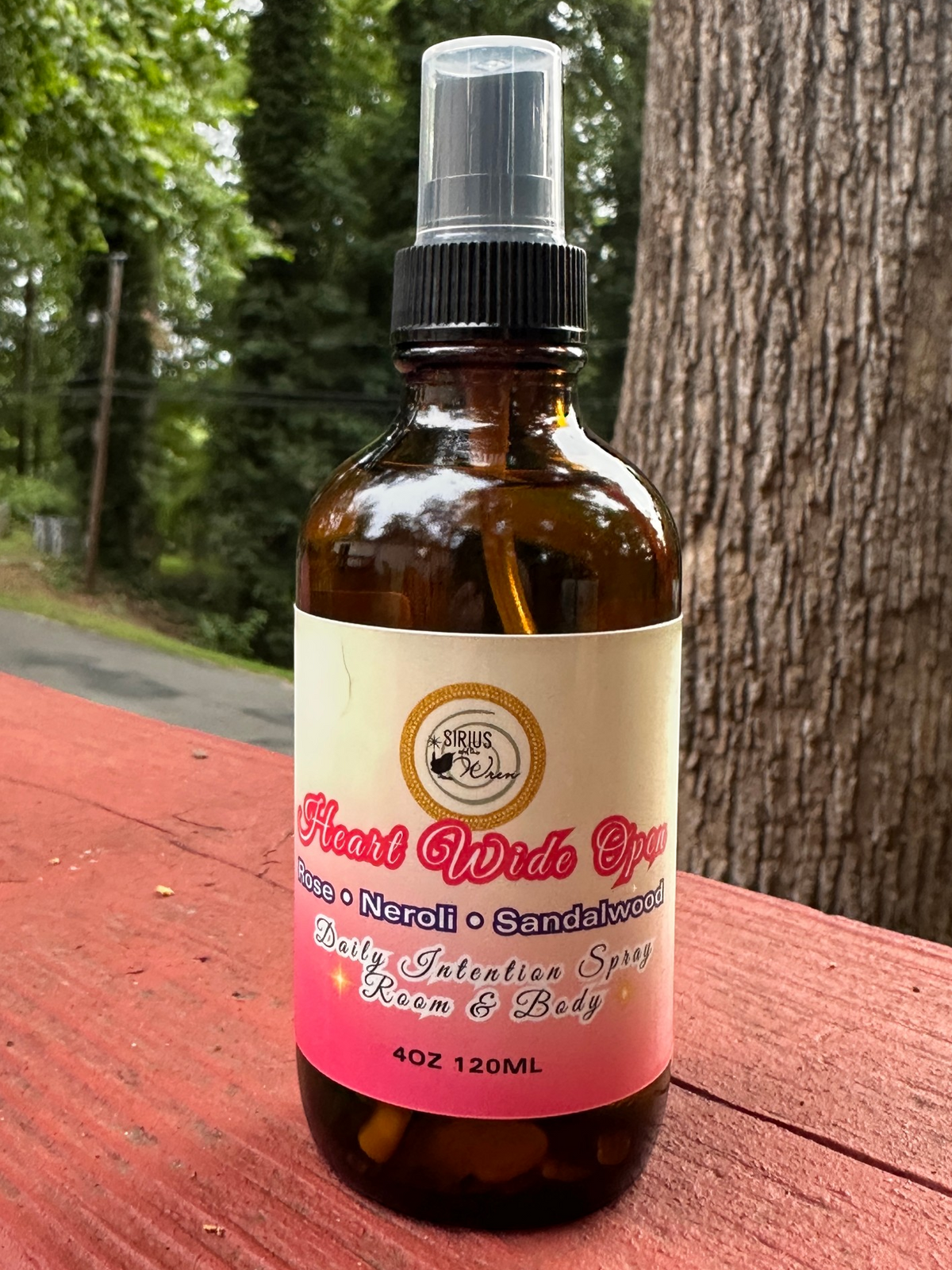 Heart Wide Open Daily Intention Spray, 4 oz, with a pink label on a wooden surface outdoors