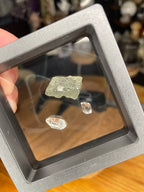 A piece of rough, olive-green Moldavite with Herkimer Diamonds - 1.15 g and two small, clear Herkimer diamonds displayed inside a suspended floating frame.
