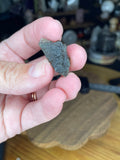 A hand holding a small, textured, dark green Moldavite with Herkimer Diamonds - 2.37 g mineral fragment.