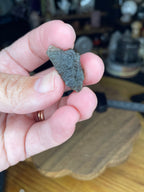 A hand holding a small, textured, dark green Moldavite with Herkimer Diamonds - 2.37 g mineral fragment.