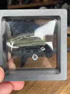 A Moldavite with Herkimer Diamonds - 3.4 g featuring a textured, olive-green stone and two small, clear, faceted crystals displayed inside a square floating frame.
