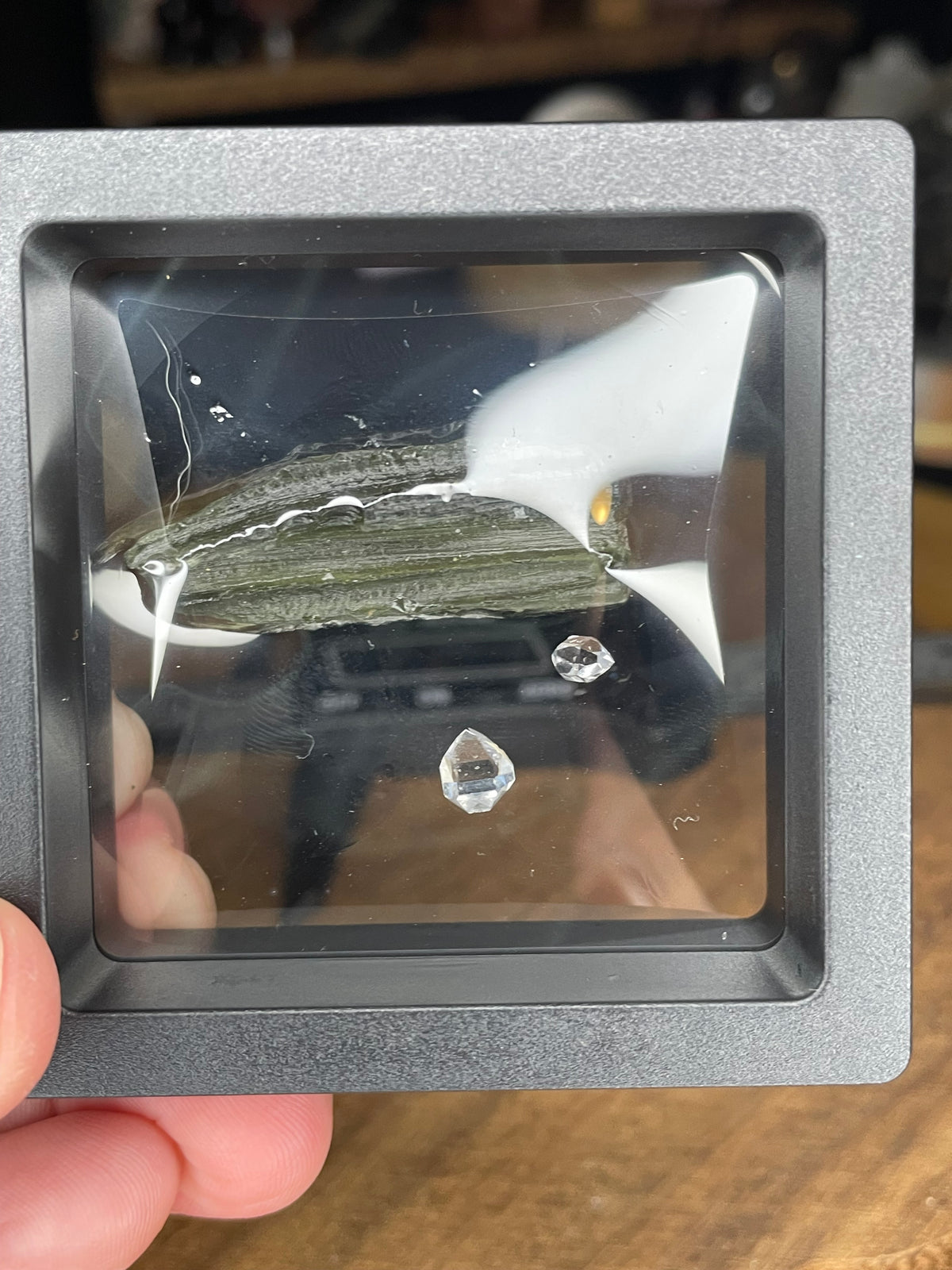 A Moldavite with Herkimer Diamonds - 3.4 g featuring a textured, olive-green stone and two small, clear, faceted crystals displayed inside a square floating frame.