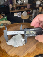 A hand holds a digital caliper measuring an Apophyllite Specimen - 108 g cluster of clear crystals resting on a wooden surface.