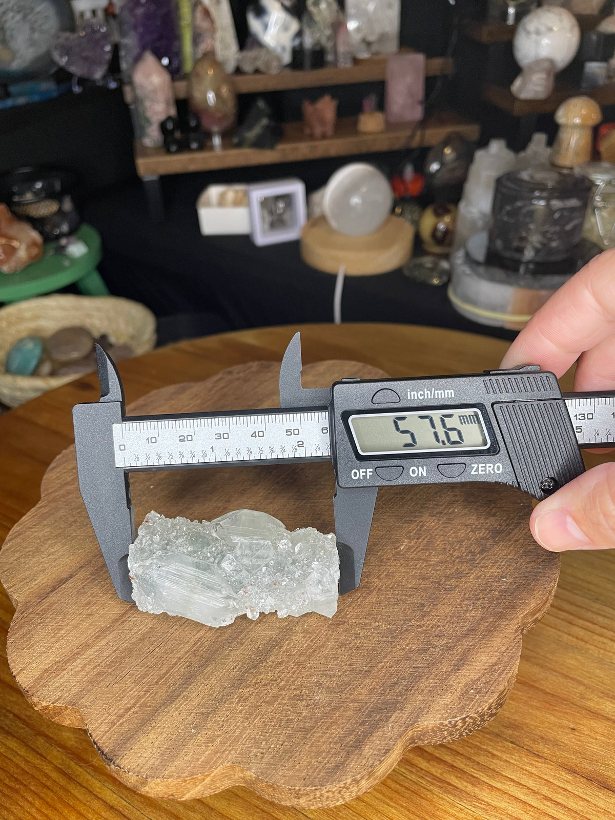 A clear, crystalline Apophyllite with Black Chalcedony cluster being measured by digital calipers showing a reading of 57.6 mm on a wooden surface.