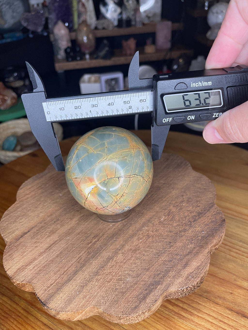 A hand holds digital calipers measuring a Creek Jasper Sphere - 63.2 mm, which has a veined pattern and a digital display reading of 63.2 millimeters.