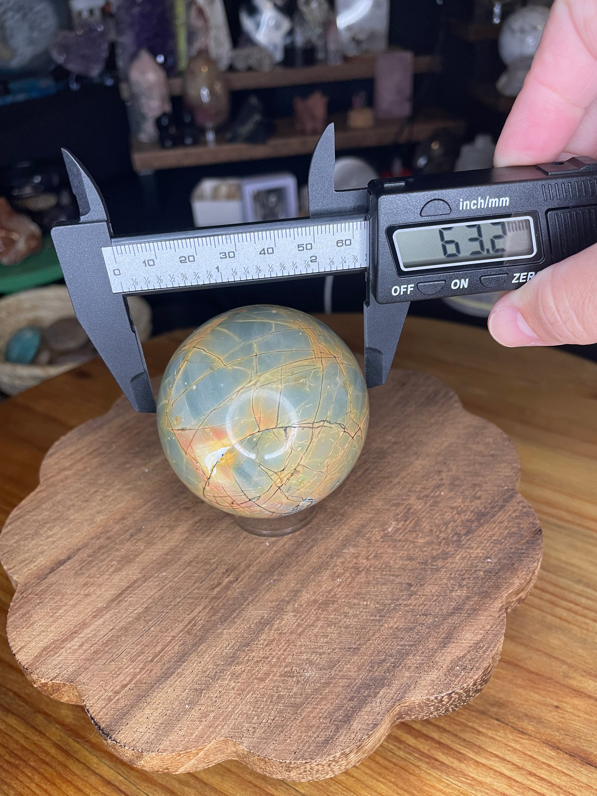 A hand holds digital calipers measuring a Creek Jasper Sphere - 63.2 mm, which has a veined pattern and a digital display reading of 63.2 millimeters.