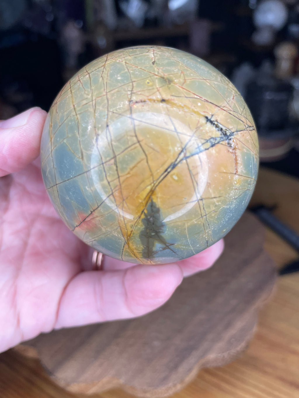 A hand holds a Creek Jasper Sphere - 63.2 mm, a polished sphere of green and tan stone with dark, web-like veining.
