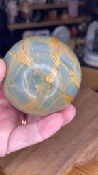 A hand holding a Creek Jasper Sphere - 63.2 mm, a smooth, spherical stone with a grey-green base and a pattern of orange and dark brown veins.