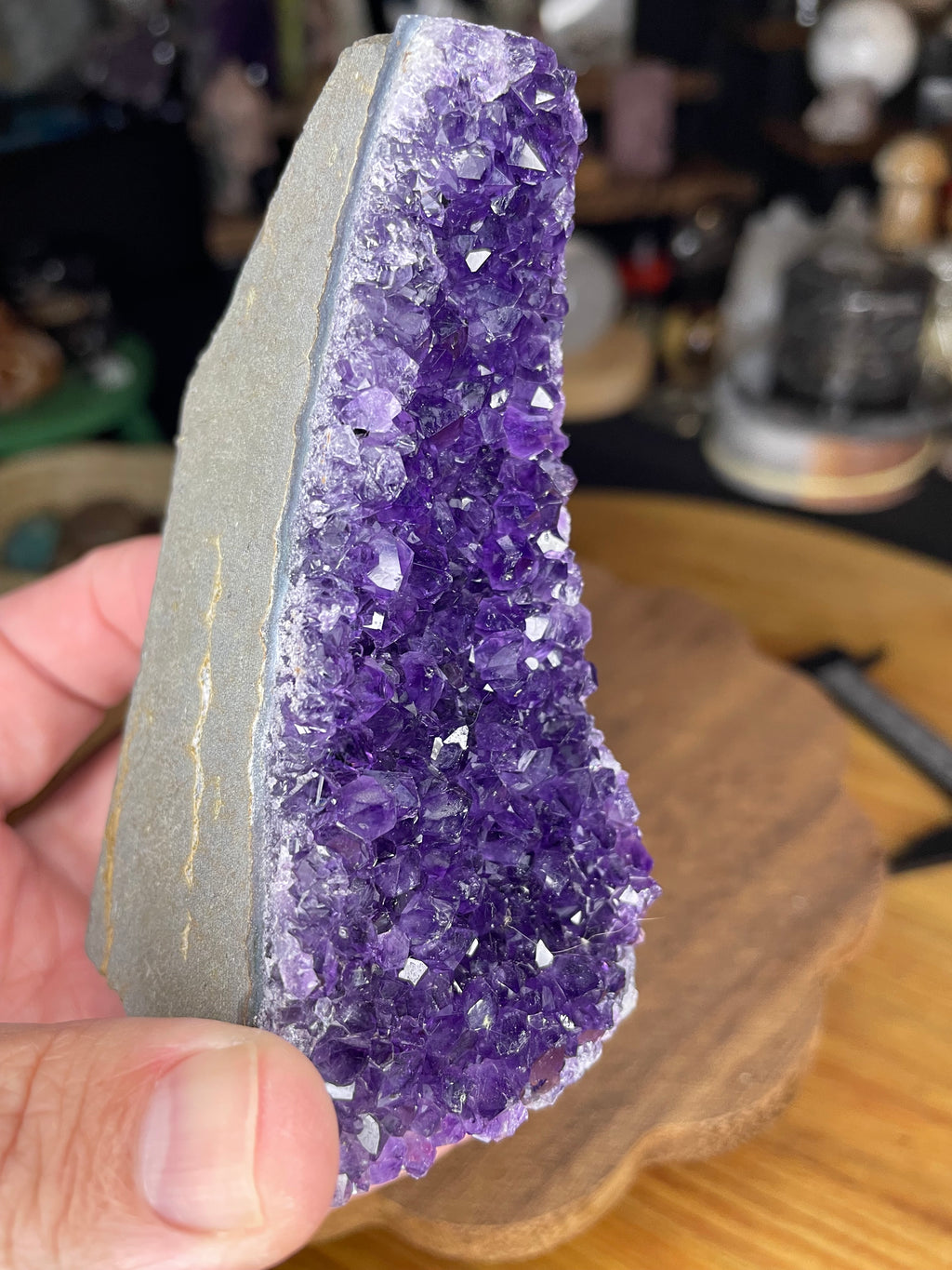 A hand holds a Brazilian Amethyst Cut Base - 304 g, a tall, wedge-shaped geode with a deep purple crystalline interior and a smooth, cut gray stone back.