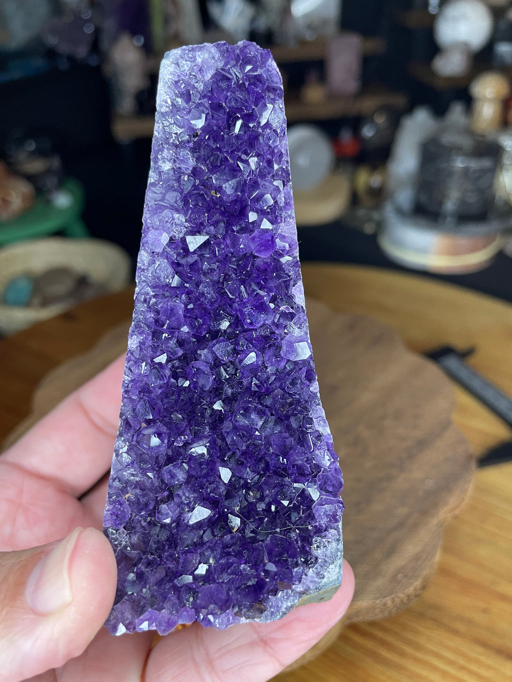 A hand holds a Brazilian Amethyst Cut Base - 304 g, a tall, triangular piece of deep purple amethyst crystal with a flat, polished base.