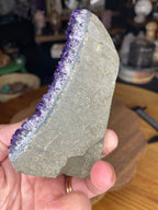 A hand holds a Brazilian Amethyst Cut Base - 304 g, featuring a flat, grey cut base and jagged purple crystal points along one edge.