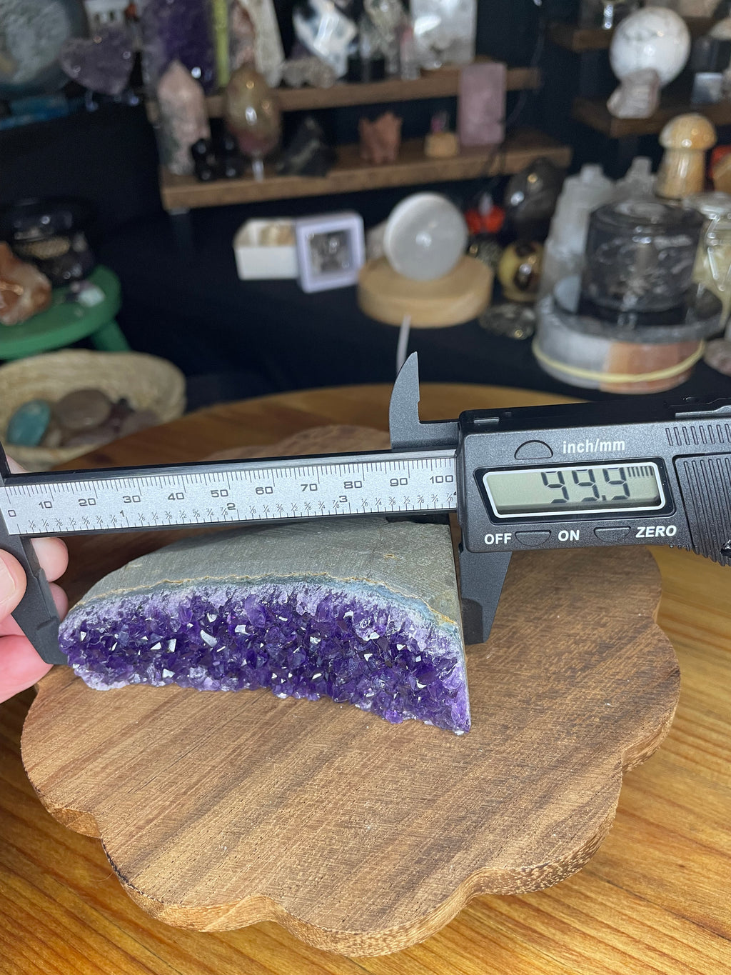 A Brazilian Amethyst Cut Base - 304 g raw geode slice with a flat bottom being measured by a digital caliper showing 99.9 millimeters.