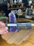 A hand uses digital calipers to measure the width of a Brazilian Amethyst Cut Base - 304 g, a purple crystal cluster on a wooden base.