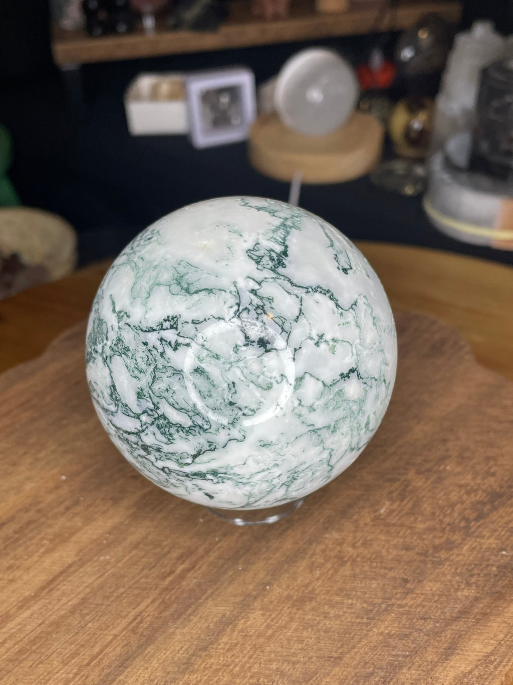 A polished Tree Agate Sphere with Druzy - 73.3 mm, featuring white and green patterns, sits on a wooden surface.