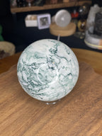 A polished Tree Agate Sphere with Druzy - 73.3 mm, featuring white and green patterns, sits on a wooden surface.