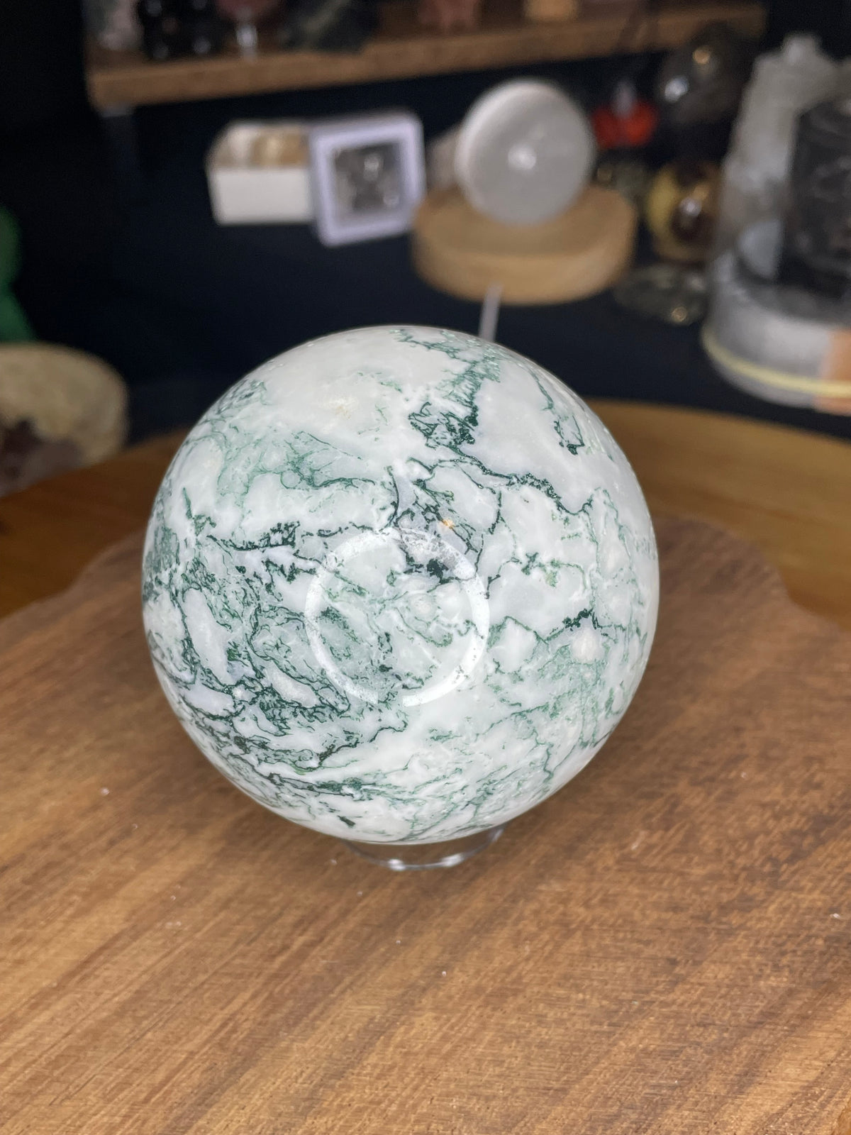 A polished Tree Agate Sphere with Druzy - 73.3 mm, featuring white and green patterns, sits on a wooden surface.