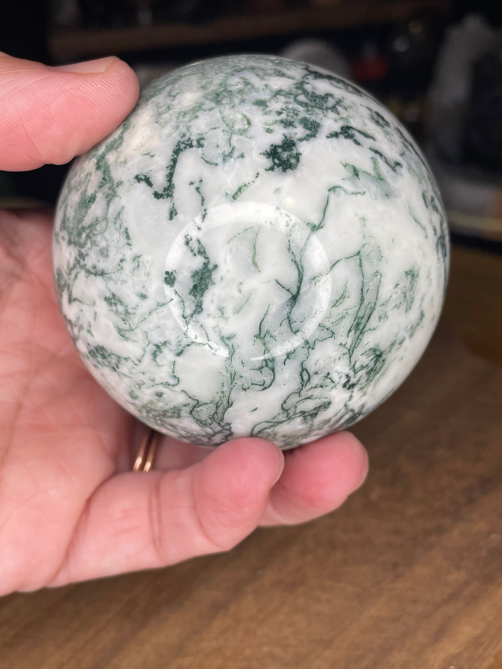 A hand holds a Tree Agate Sphere with Druzy - 73.3 mm, a polished sphere of white stone with green, moss-like inclusions.