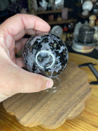 A hand holds a 66.7 mm Indigo Gabbro (Mystic Merlinite) Sphere, featuring a polished, patterned surface with dark grey, black, and metallic flecks.