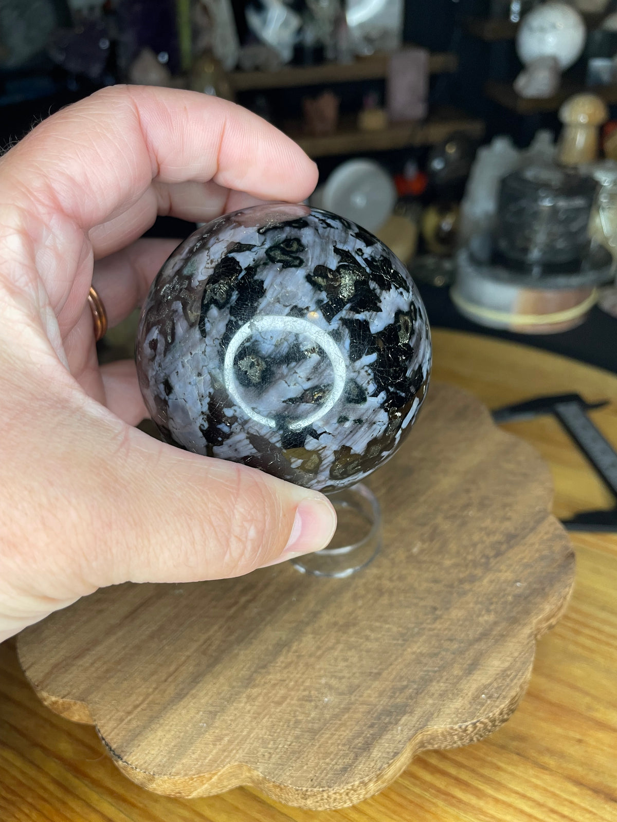 A hand holds a 66.7 mm Indigo Gabbro (Mystic Merlinite) Sphere, featuring a polished, patterned surface with dark grey, black, and metallic flecks.