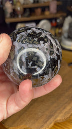 A hand holds a 66.7 mm Indigo Gabbro (Mystic Merlinite) Sphere patterned with dark, marbled swirls and metallic gold flecks.