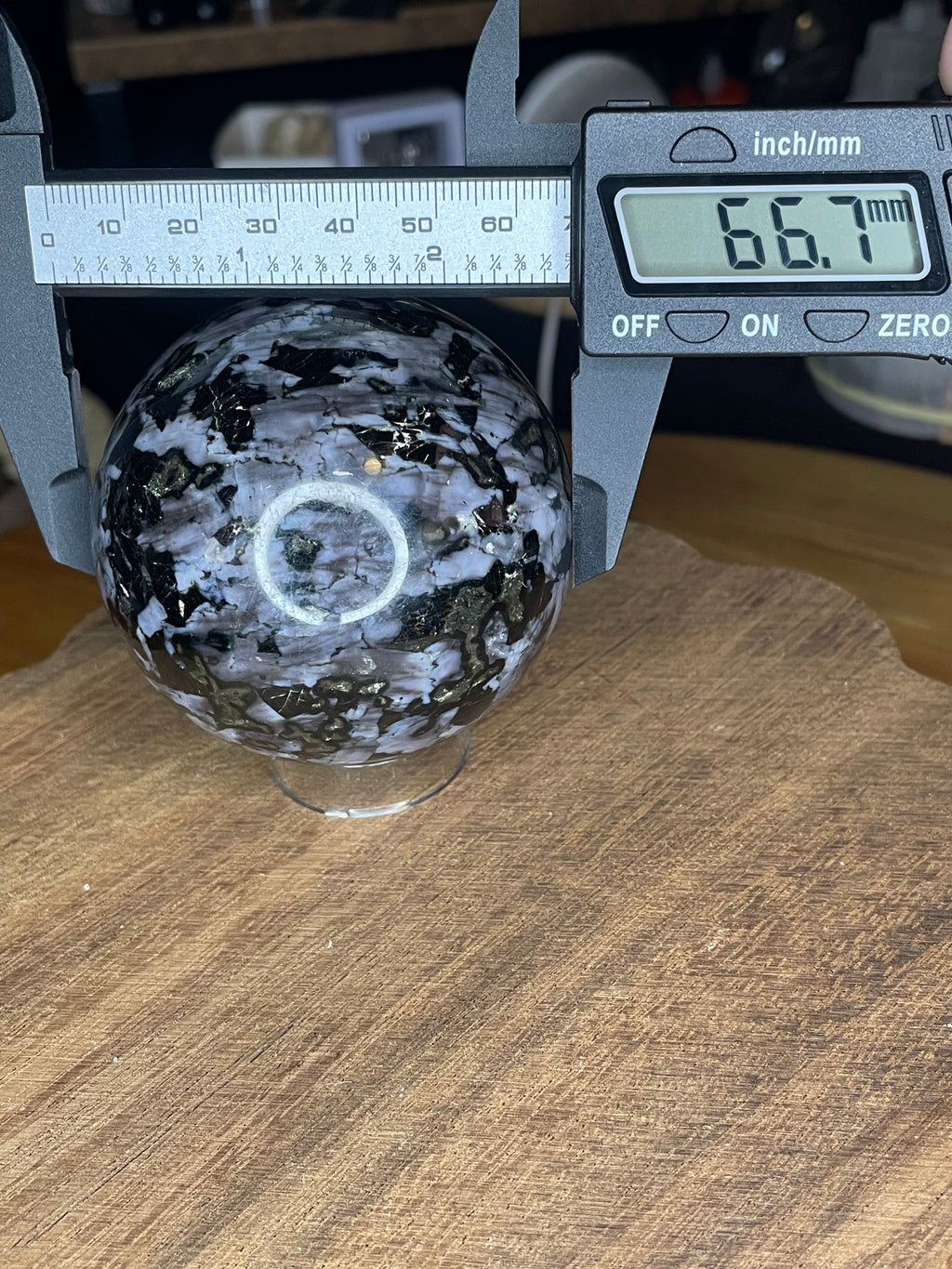 An Indigo Gabbro (Mystic Merlinite) Sphere - 66.7 mm, shown as a black and white speckled stone sphere measuring 66.7 millimeters on a digital caliper.