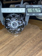 An Indigo Gabbro (Mystic Merlinite) Sphere - 66.7 mm, shown as a black and white speckled stone sphere measuring 66.7 millimeters on a digital caliper.