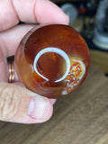 A hand holds a Carnelian Sphere - 48.2 mm, a polished, reddish-orange gemstone with white inclusions and a circular light reflection.