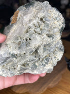 A hand holds a Brazilian Blue Tara Quartz Cluster - 600 g, featuring a jagged cluster of translucent, pale blue and white crystals.
