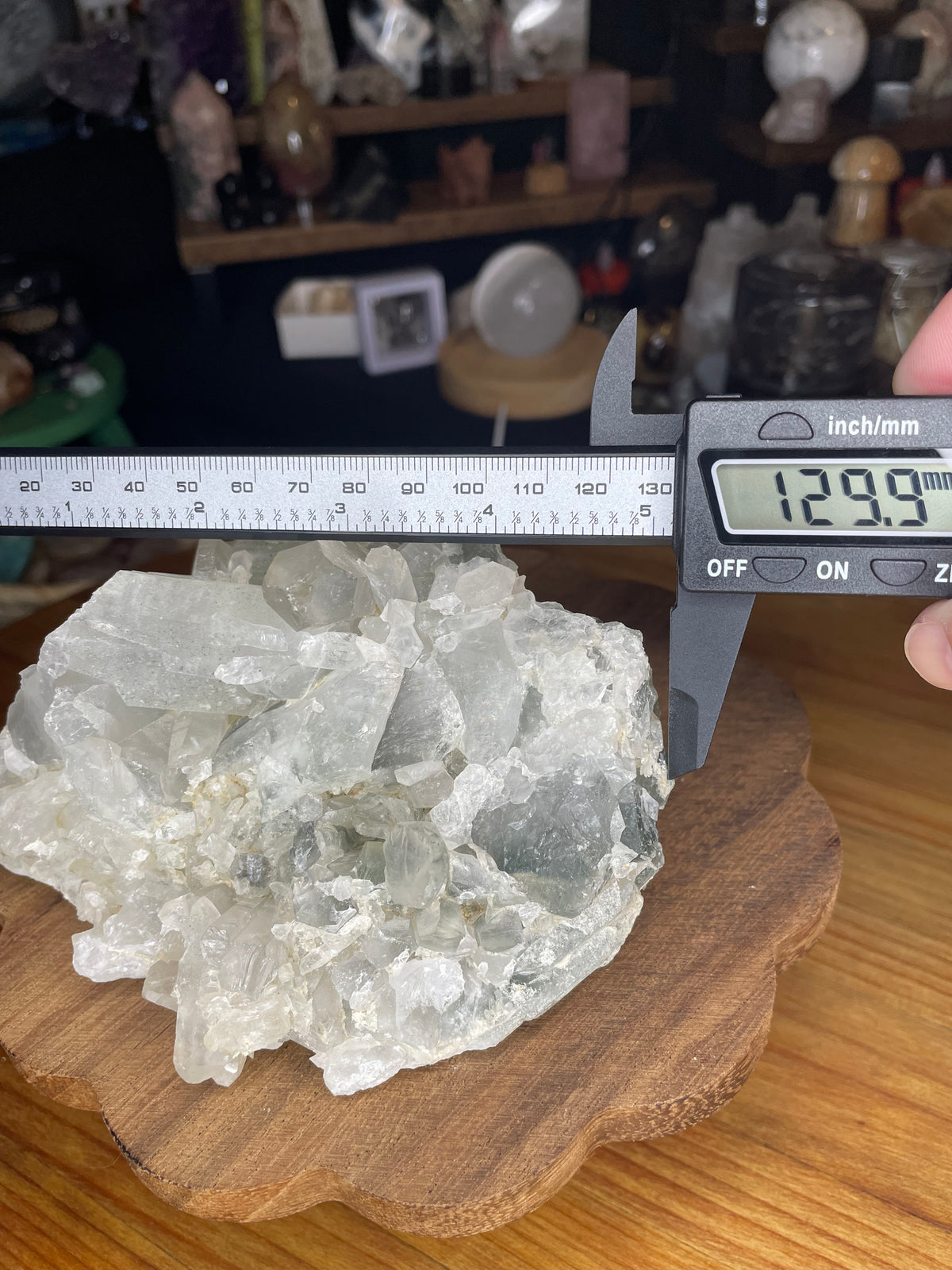 A Brazilian Blue Tara Quartz Cluster being measured with a digital caliper that reads 129.9 mm, showing its translucent, light-colored crystals.