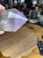 A hand holding a 428 g Chunky Rainbow Fluorite Tower - UV Reactive - from Sirius and the Wren, featuring a polished crystal with distinct purple and clear layers.
