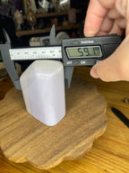 A hand uses digital calipers to measure a Rare Lavender Yttrium Fluorite Freeform - 414 g specimen resting on a wooden stand.