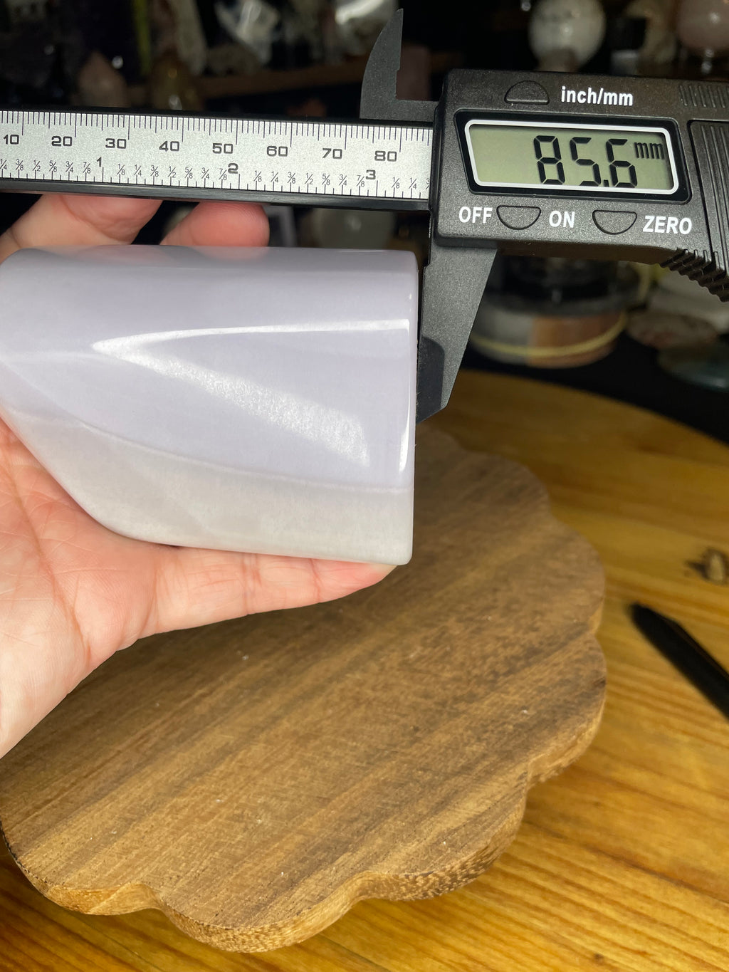 A hand holds a Rare Lavender Yttrium Fluorite Freeform being measured by a digital caliper showing 85.6 millimeters.