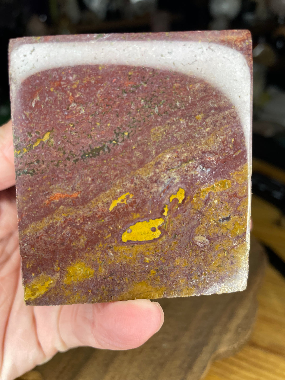 A hand holds an Ocean Jasper Floating Cube - 18.5 oz, a cube-shaped stone with swirling patterns of red, yellow, and white.