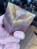 A hand holding an Ocean Jasper Floating Cube - 18.5 oz, a stone shaped like a cube with swirling bands of red, yellow, and brown.