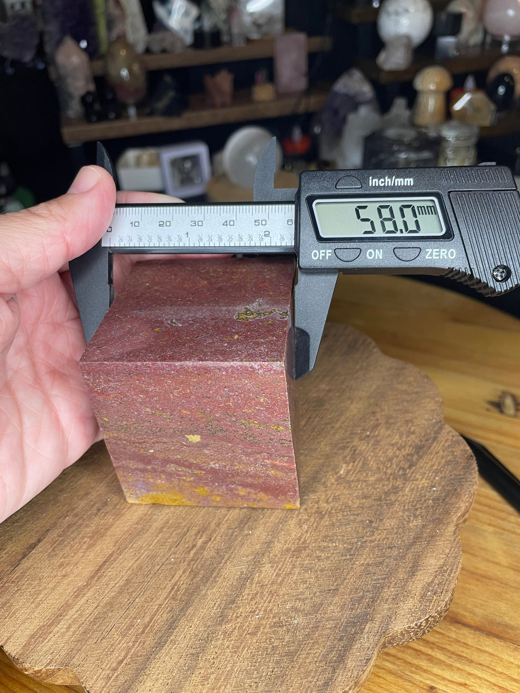 A hand holds a digital caliper measuring 58.0 mm across a red and yellow Ocean Jasper Floating Cube - 18.5 oz.