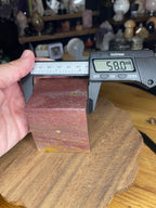 A hand holds a digital caliper measuring 58.0 mm across a red and yellow Ocean Jasper Floating Cube - 18.5 oz.