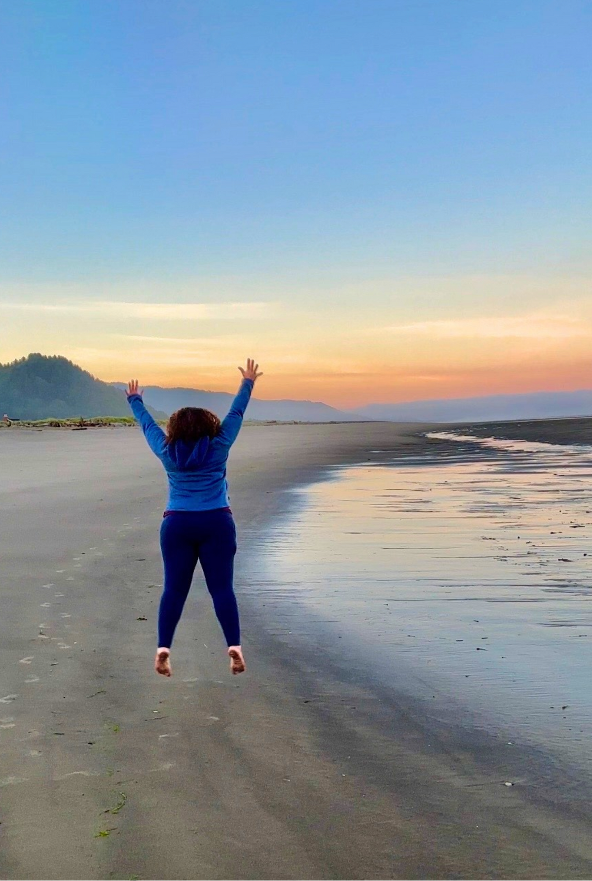 A woman in a blue long-sleeve top and dark leggings jumps barefoot on a wide sandy beach at sunset, with her arms raised above her head. The background features a calm ocean shoreline, wet sand reflecting the sky, and distant mountains under a gradient sunset sky.